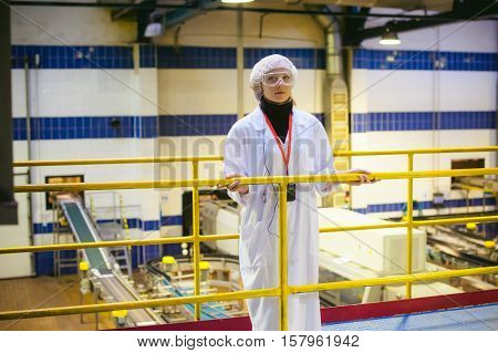 Female Worker On The Production Line Of Beer Factory. Portrait Of A Woman In A White Robe, Standing