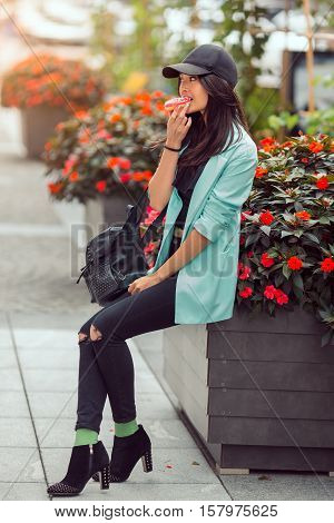 Asian beautiful woman eating a donut sitting near bed of flowers on street, fast food concept outdoors. Busy businessman have no time.