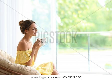 Young Woman With Cup Of Aromatic Coffee Sitting In Papasan Chair Near Window At Home
