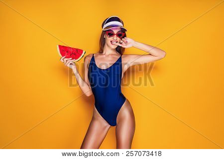 Beautiful Young Lady In Swimwear Biting Finger And Holding Piece Of Fresh Watermelon While Standing 