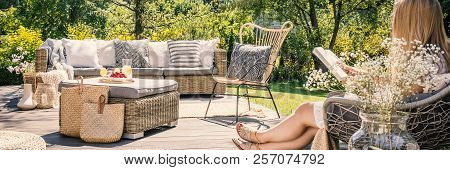 Woman Reading Book On Patio With Rattan Table, Chair And Sofa In The Garden During Summer. Real Phot