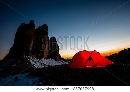 Night bivouac at Tre Cime di Lavaredo, milion star hotel under night sky, red illuminated tent on pass in Dolomites, Italy.