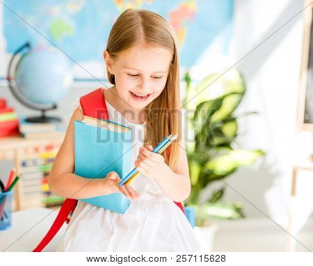 Little smiling blond girl standing near blackbord with red backpack and holding a notebook with pencils in the sunshine school classroom