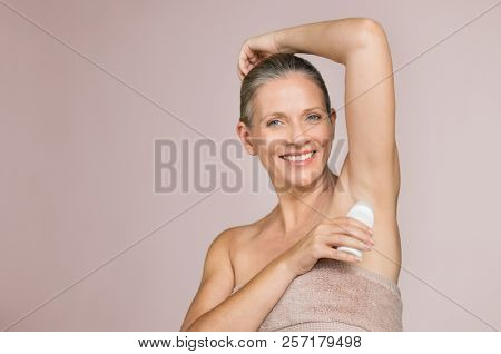 Smiling woman applying deodorant in armpit isolated on grey background with copy space. Beautiful senior woman putting antiperspirant stick after shower and looking at camera.