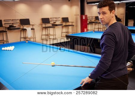 Man in dark blue shirt stands half-turned at pool table with balls pyramid in club.