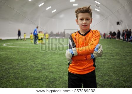 Boy in sports uniform with goalkeeper gloves on hands standing on indoor football field.