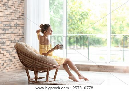 Young Woman With Cup Of Aromatic Coffee Sitting In Papasan Chair Near Window At Home