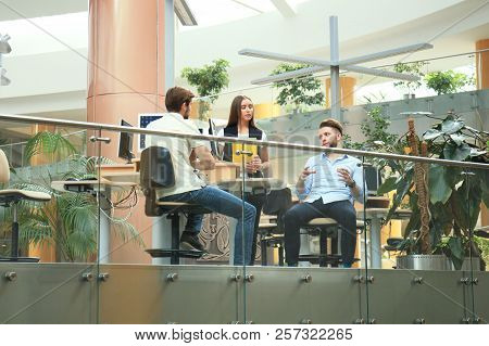 Group Of Young People In Casual Wear Sitting At The Office Desk And Discussing Something While Looki