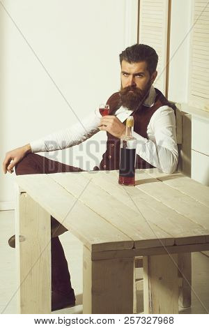 Hipster Drinking Red Wine With Bottle On Wooden Table. Brutal Man With Long Beard And Moustache Indo