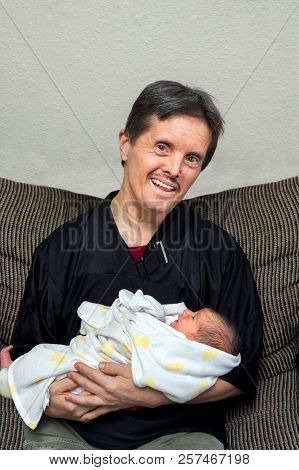A Happy Man With Downs Syndrome And Fake Teeth Holds A Newborn Baby.  He Has A Big Smile As He Looks
