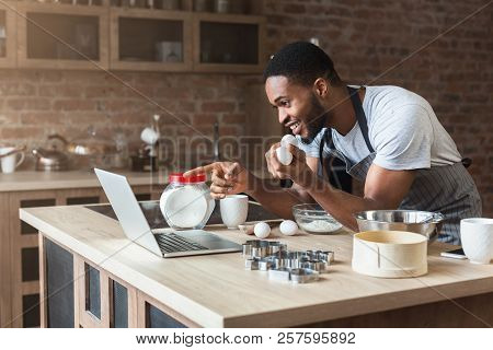 Excited Black Man Baking Pastry And Looking Recipe On Laptop In Loft Kitchen