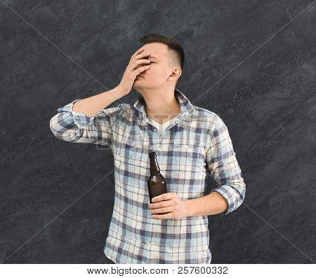 Young Guy Alcoholic With Headache Holding Bottle Of Beer, Suffering From Hangover, Dark Studio Backg