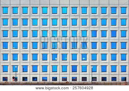 A Small Inconspicuous Human Walks Along A Tall Building. Many Windows On The Facade Of The Building.