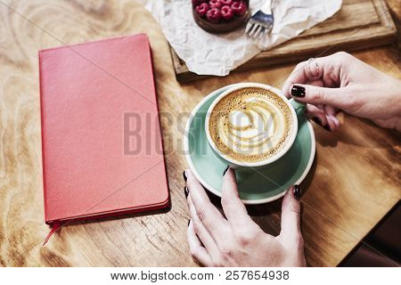 Cup Of Coffee Latte And Red Book On Wooden Table Or Background In Woman Hands From Above.