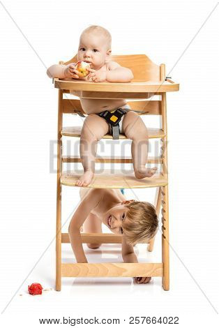 Baby Girl Sitting In High Chair And Eating Apple, Preschool Boy Hiding Under Footplate. Siblings Pla