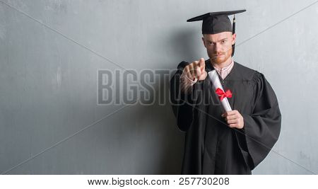 Young redhead man over grey grunge wall wearing graduate uniform holding degree pointing with finger to the camera and to you, hand sign, positive and confident gesture from the front