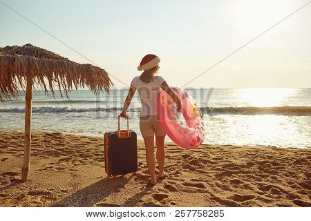 A Girl In A Santa Claus Hat With A Suitcase On The Beach On Chri