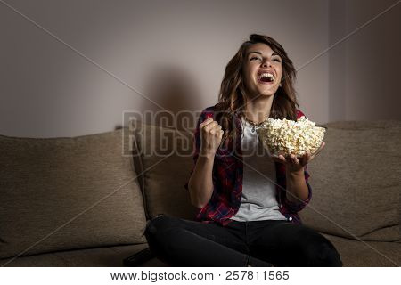 Beautiful Young Woman Sitting In The Dark On A Living Room Couch, Having Fun Watching A Football Mat
