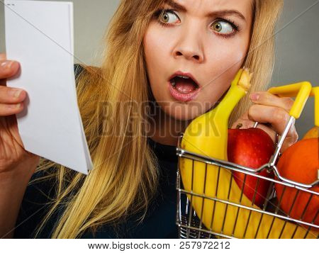 Shocked Woman Holding Shopping Basket With Fruits Looking At Bill Receipt Being Scared Of Huge Price