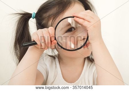 Little Girl Plays A Detective. A Child Examines Something Through A Magnifying Glass. A Girl With Tw