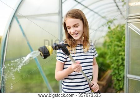 Adorable Young Girl Watering Variuos Vegetables In A Greenhouse On Sunny Summer Day. Gardening Activ