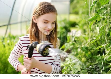 Adorable Young Girl Watering Variuos Vegetables In A Greenhouse On Sunny Summer Day. Gardening Activ