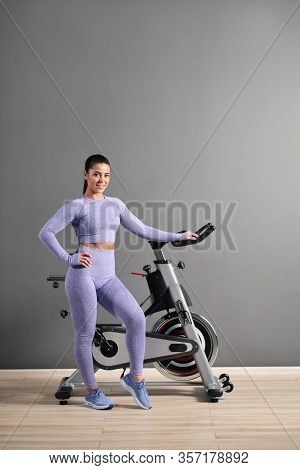 A Beautiful Athletic Young Brunette Woman In Sportswear Trains On A Sycle In The Gym Against The Gra