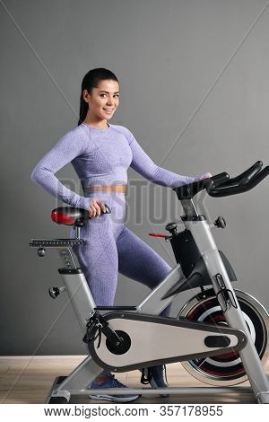 A Beautiful Athletic Young Brunette Woman In Sportswear Trains On A Sycle In The Gym Against The Gra