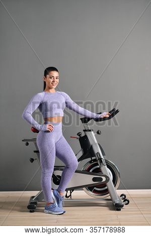 A Beautiful Athletic Young Brunette Woman In Sportswear Trains On A Sycle In The Gym Against The Gra
