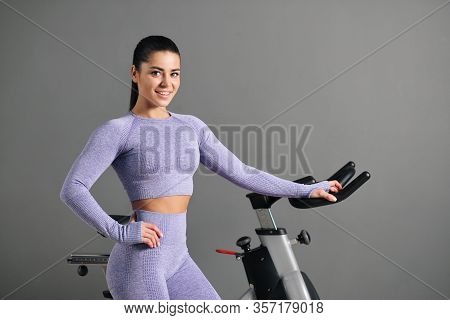 A Beautiful Athletic Young Brunette Woman In Sportswear Trains On A Sycle In The Gym Against The Gra
