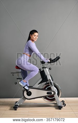 A Beautiful Athletic Young Brunette Woman In Sportswear Trains On A Sycle In The Gym Against The Gra