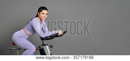 A Beautiful Athletic Young Brunette Woman In Sportswear Trains On A Sycle In The Gym Against The Gra