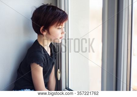 Sad Little Girl Looking Out Window. Child During Quarantine Because Of Pandemic Of Coronavirus Stari