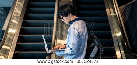 Asian Man With Glasses And Bag Holding Laptop Computer Standing In Front Of Escalator In Publing Bui