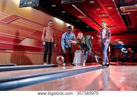 Pretty young blond female throwing bowling ball on track while standing among her intercultural friends during play