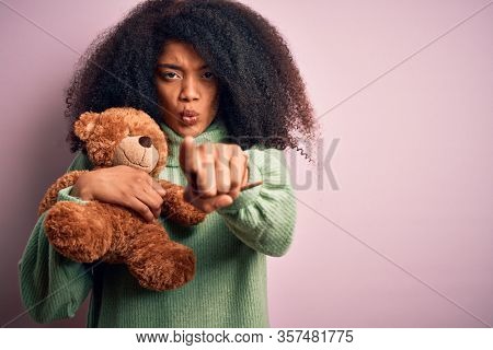 Young african american woman with afro hair hugging teddy bear over pink background pointing with finger to the camera and to you, hand sign, positive and confident gesture from the front