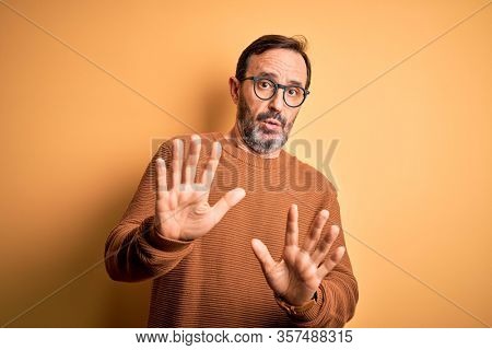 Middle age hoary man wearing brown sweater and glasses over isolated yellow background Moving away hands palms showing refusal and denial with afraid and disgusting expression. Stop and forbidden.