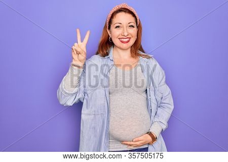 Young beautiful redhead pregnant woman expecting baby over isolated purple background showing and pointing up with fingers number two while smiling confident and happy.