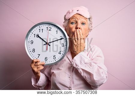 Senior beautiful woman wearing sleep mask holding big clock over isolated pink background cover mouth with hand shocked with shame for mistake, expression of fear, scared in silence, secret concept
