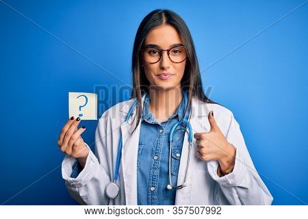 Young beautiful brunette doctor woman holding paper with question mark symbol message happy with big smile doing ok sign, thumb up with fingers, excellent sign