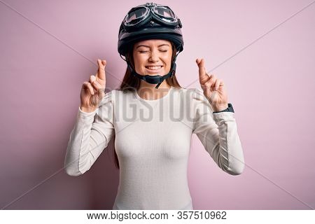 Young beautiful motorcyclist woman with blue eyes wearing moto helmet over pink background gesturing finger crossed smiling with hope and eyes closed. Luck and superstitious concept.