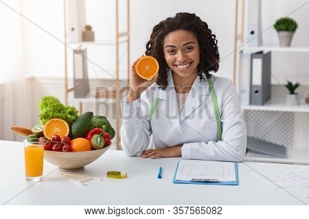Modern Dietologist African Woman Sitting At Workplace And Recommending Fresh Oranges