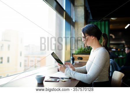 Hipster Girl Skilled E-book Writer Reading Information On Website Via Portable Touch Pad While Sitti