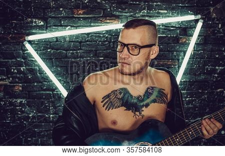 Tattooed Man Playing Guitar Near Illuminated Wall. Stylish Guy With Bird Tattoo On Chest Looking Awa