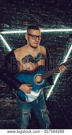 Tattooed Man Playing Guitar Near Illuminated Wall. Stylish Guy With Bird Tattoo On Chest Looking Awa