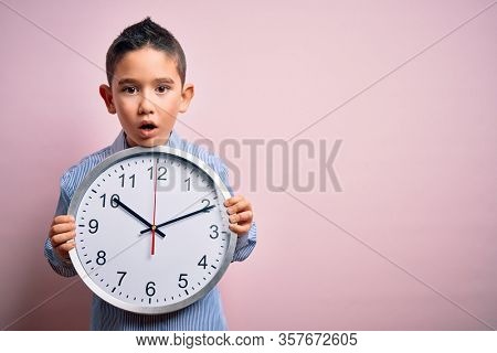 Young little boy kid holding big minute clock over isolated pink background scared in shock with a surprise face, afraid and excited with fear expression