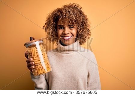 Young african american curly woman holding jar with Italian dry pasta macaroni with a happy face standing and smiling with a confident smile showing teeth