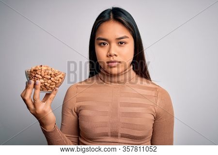 Young asian girl holding bowl of healthy peanuts eating as snack for diet over white background with a confident expression on smart face thinking serious