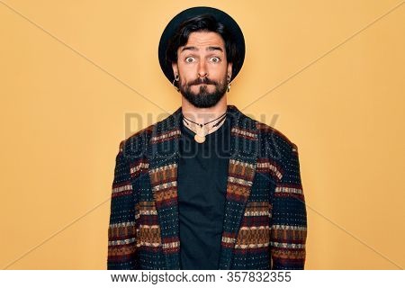 Young handsome hispanic bohemian man wearing hippie style and boho hat puffing cheeks with funny face. Mouth inflated with air, crazy expression.