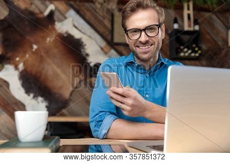 casual man wearing glasses sitting at desk and texting a message on phone while looking at camera happy at the coffeeshop 